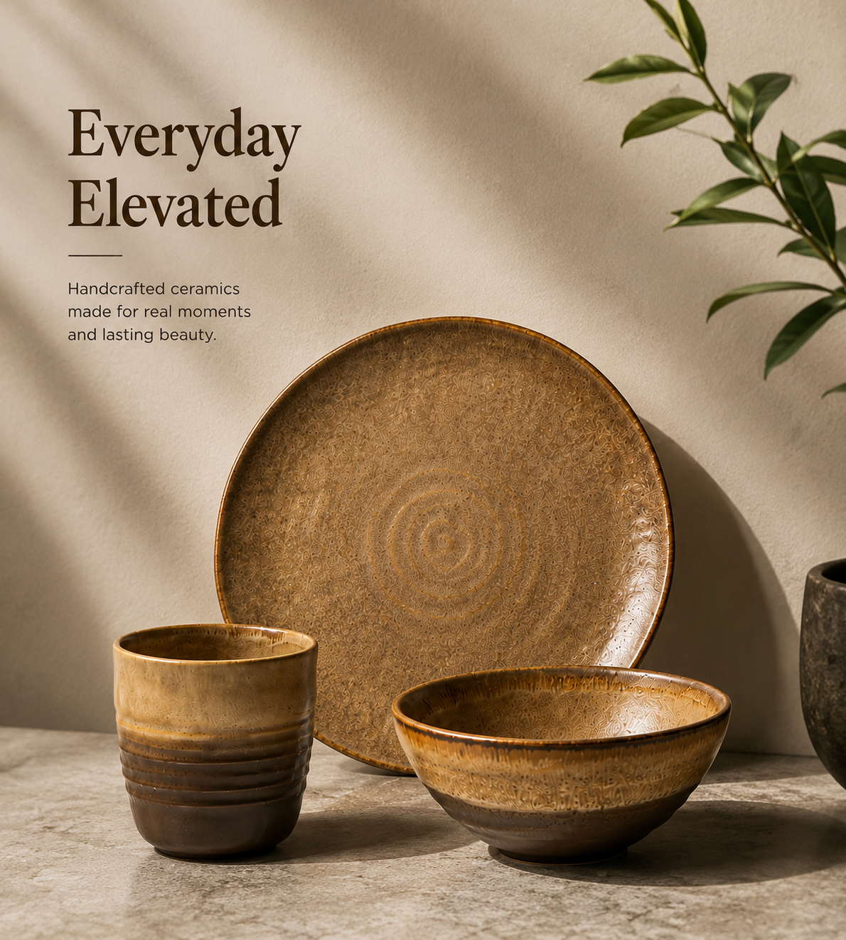 Three handcrafted ceramic tableware pieces in earthy brown and tan tones, including a large plate, a shallow bowl, and a small cup, arranged on a stone-like surface against a beige wall with soft shadows and a plant leaf in the corner. 